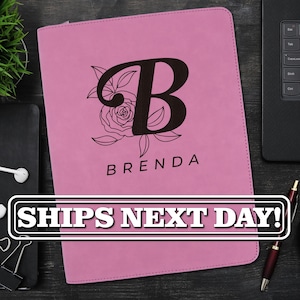May include: A pink rectangular portfolio folder featuring a large black letter B monogram overlaid on a floral line art design, with the name BRENDA printed in black capital letters below the monogram on the front cover surface.