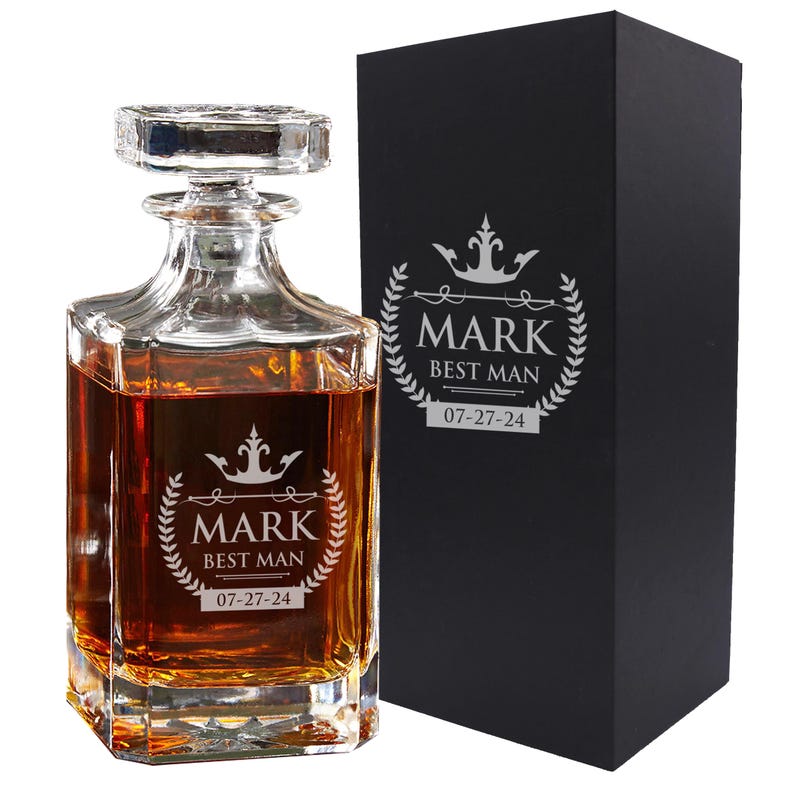 Peut inclure: Une carafe en verre transparent remplie d'un liquide ambr&eacute;, avec un bouchon carr&eacute; et une gravure repr&eacute;sentant une couronne, une couronne de laurier, le nom MARK, le texte BEST MAN et la date 07-27-24, &agrave; c&ocirc;t&eacute; d'une bo&icirc;te cadeau noire.