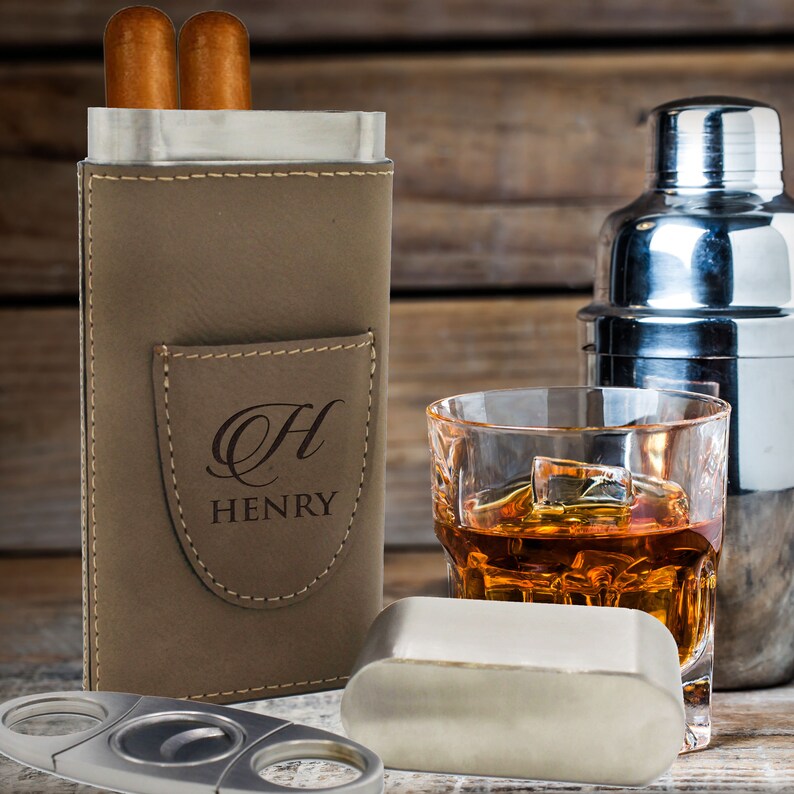May include: A taupe leather cigar case with a front pocket featuring the engraved letter H and the name HENRY. The case holds two cigars and is accompanied by a silver metal cigar cutter and a separate silver metal cap on a wooden surface.