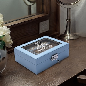 May include: A light blue rectangular jewelry box with a clear glass lid featuring a white printed monogram with the letter M and the name MADISON. The box includes a silver-toned metal latch with a small key inserted into the lock mechanism.