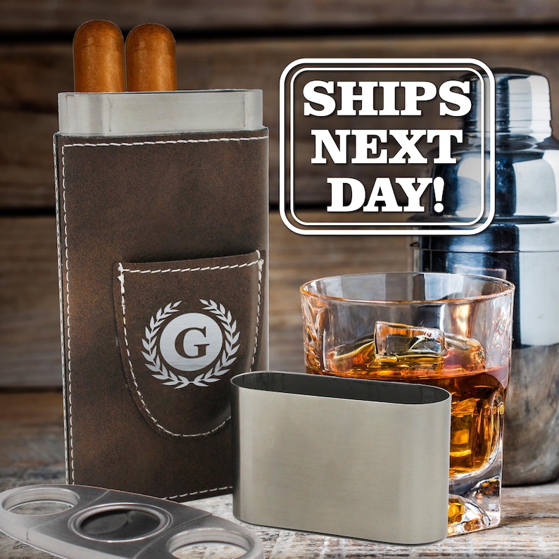 May include: A brown leather cigar case with white stitching and a letter G inside a laurel wreath emblem, containing two cigars. Includes a separate stainless steel cigar cutter, a metal sleeve, and a glass of amber liquid with ice cubes.