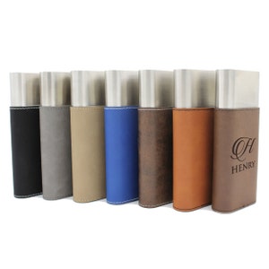 May include: A row of seven rectangular metal flasks with leather-like sleeves in black, gray, tan, blue, dark brown, light brown, and a tan sleeve with the engraved text H HENRY. Each flask features a brushed silver metal top section.