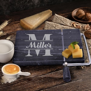 May include: Black marble cheese board with a wire cheese cutter and a personalized engraved letter "M" with the name "Miller" underneath. The board is on a wooden surface with a block of cheese and a small bowl of milk.