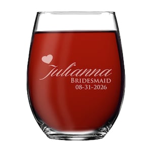 May include: A clear stemless wine glass filled with red liquid, featuring white etched text that reads Julianna, BRIDESMAID, and 08-31-2026, accompanied by a small white heart icon positioned above the name on the front of the glass.
