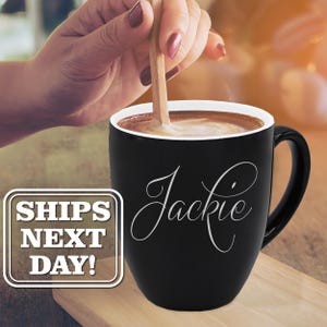 May include: A black ceramic mug with a white interior and a curved handle, featuring the name Jackie in white cursive script on the front. A rectangular white box with a brown border contains the text SHIPS NEXT DAY! in bold white letters.