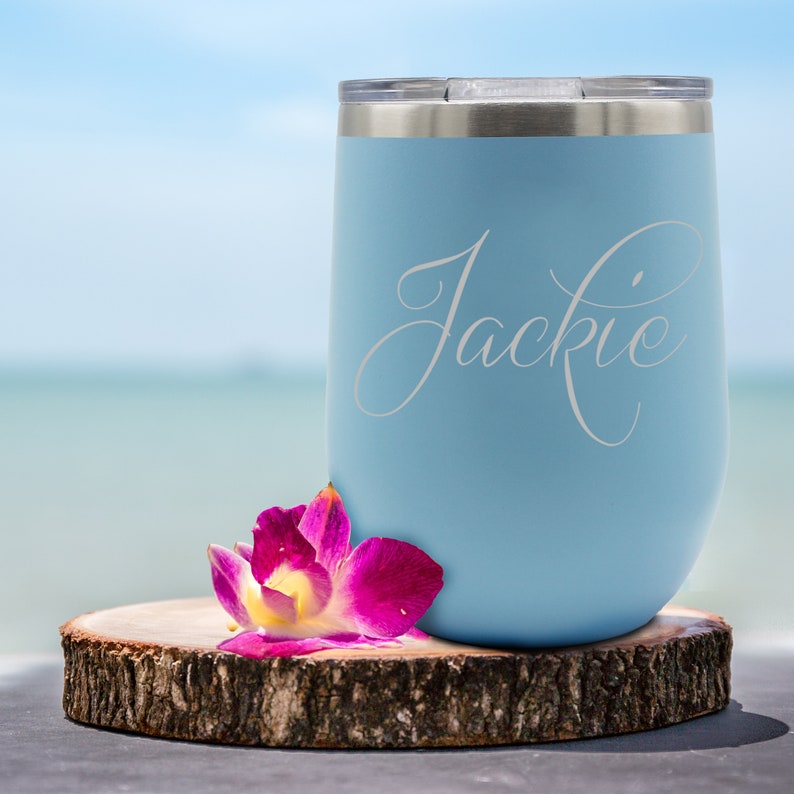 May include: A light blue insulated tumbler with the name "Jackie" in a cursive font. The tumbler sits on a wooden slice with a pink and yellow orchid. The background is a blurred beach scene.