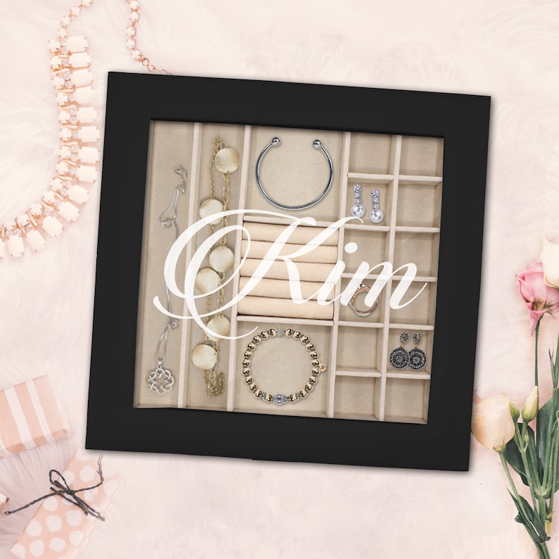 May include: A black square jewelry display case with a glass front featuring the white script text Kim. The interior is lined with beige velvet and divided into multiple compartments containing a silver cuff, a gold beaded bracelet, and earrings.