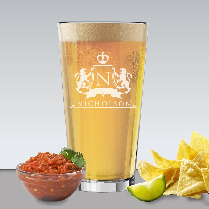 May include: A pint glass filled with beer, featuring a white crest design with the name "Nicholson." The glass is accompanied by a bowl of salsa, tortilla chips, and a lime wedge, suggesting a casual gathering.