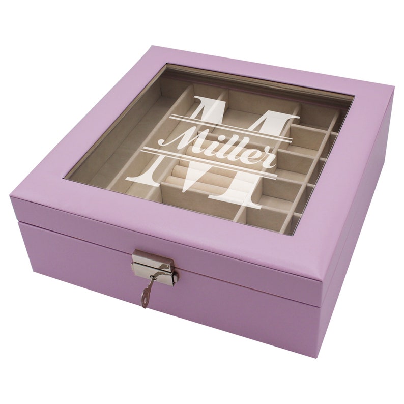 May include: A square lavender jewelry box with a clear glass lid featuring a white monogram letter M and the name Miller printed in the center. The box includes a silver metal latch with a small key and a beige velvet interior with multiple compartments.