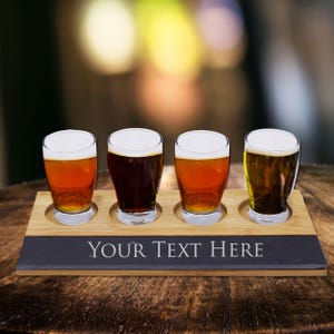 May include: A wooden beer flight set with four glasses filled with different beers. The set includes a bamboo tray and a slate base with the text "YOUR TEXT HERE" in white. The beers vary in color from light amber to dark brown.