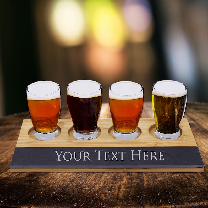 May include: A wooden beer flight set with four glasses filled with different beers. The set includes a bamboo tray and a slate base with the text "YOUR TEXT HERE" in white. The beers vary in color from light amber to dark brown.