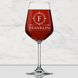 May include: A clear glass wine glass filled with deep red liquid. The glass features a white etched design consisting of a circular filigree frame surrounding the letter F, with the name FRANKLIN printed in white capital letters underneath.