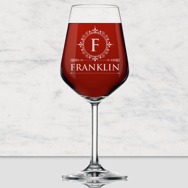 May include: A clear glass wine glass filled with deep red liquid. The glass features a white etched design consisting of a circular filigree frame surrounding the letter F, with the name FRANKLIN printed in white capital letters underneath.