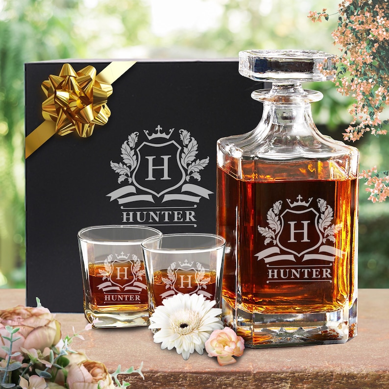 Peut inclure: Une carafe en verre transparent et deux verres assortis remplis de liquide ambr&eacute;, chacun grav&eacute; d'un blason avec la lettre H et le nom HUNTER. Une bo&icirc;te cadeau noire avec un ruban et un n&oelig;ud dor&eacute;s se trouve derri&egrave;re le set de verrerie.