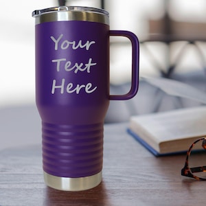 May include: A purple insulated tumbler with a handle and a silver rim and base. The text "Your Text Here" is printed in white on the front. The tumbler is on a wooden surface.