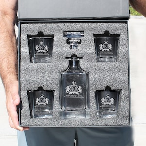Peut inclure: Une carafe en verre transparent et quatre verres assortis dans une bo&icirc;te doubl&eacute;e de mousse noire. Chaque pi&egrave;ce pr&eacute;sente un blason grav&eacute; avec une couronne, la lettre N et le nom NICHOLSON. La carafe inclut un bouchon en verre et les verres sont carr&eacute;s.