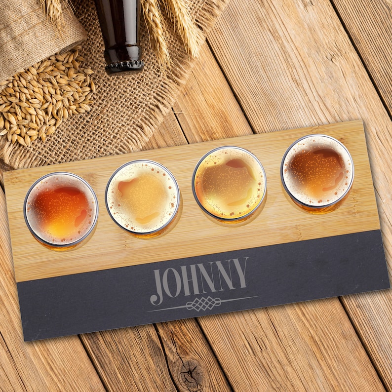 May include: A wooden beer flight board with four glasses of beer, each with a different color and foam. The board has a dark gray section with the name "JOHNNY" engraved. Barley and a beer bottle are in the background.