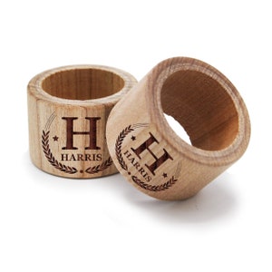 May include: Two wooden napkin rings with a personalised design. Each ring features a wreath with a star and the letter "H" in the centre. The text "HARRIS" is engraved below the letter.