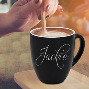 May include: A black coffee mug with the name "Jackie" in a white script font. The mug is filled with a creamy coffee drink, and a wooden spoon is stirring the contents. The mug sits on a wooden surface.