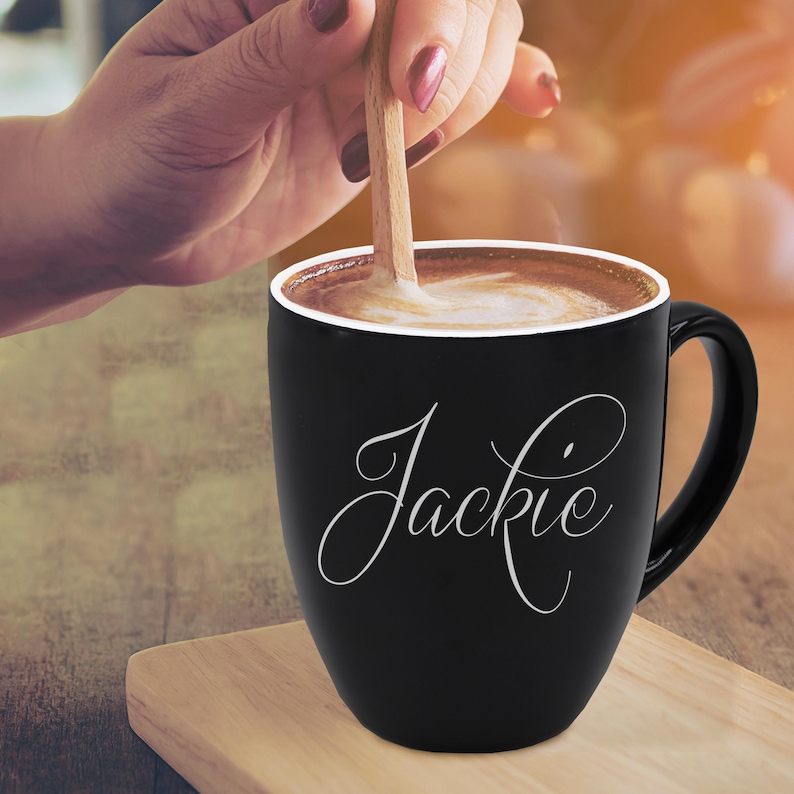 May include: A black coffee mug with the name "Jackie" in a white script font. The mug is filled with a creamy coffee drink, and a wooden spoon is stirring the contents. The mug sits on a wooden surface.