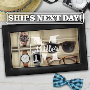 May include: A black rectangular storage box with a glass lid featuring the white text M Miller. The interior is divided into compartments containing two wristwatches, a smartwatch, and two pairs of glasses. The box has a beige velvet lining.