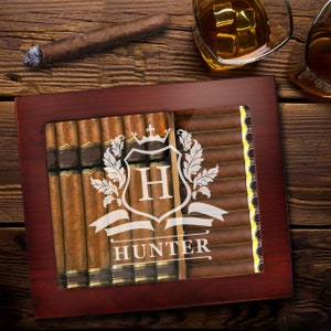 May include: A dark wood cigar humidor with a glass top featuring a white etched design of a crest with the letter H and the name HUNTER below it. The humidor is filled with rows of brown cigars, visible through the clear glass window panel.
