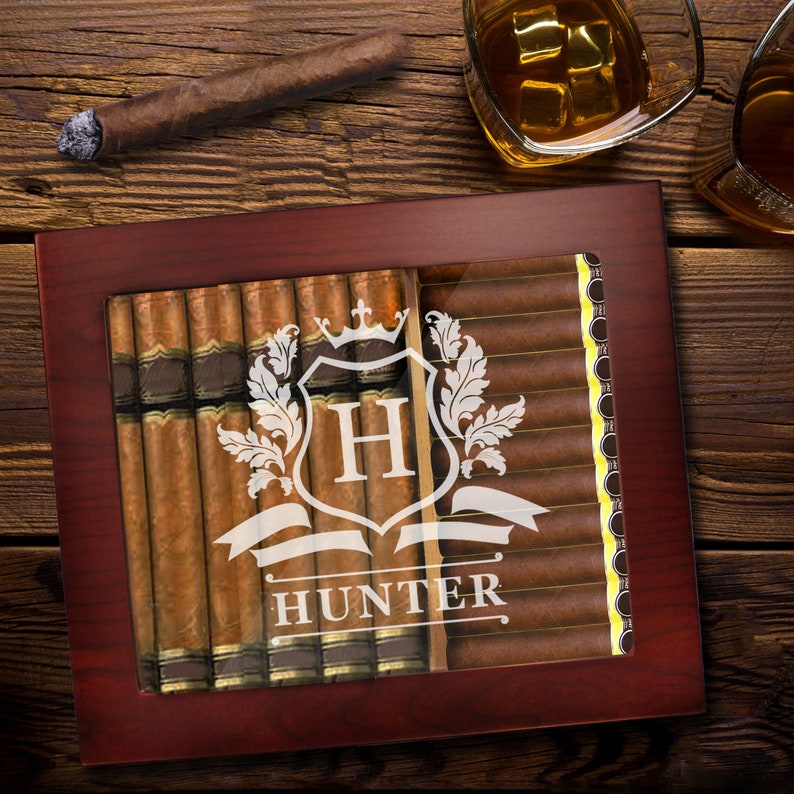 May include: A dark wood cigar humidor with a glass top featuring a white etched design of a crest with the letter H and the name HUNTER below it. The humidor is filled with rows of brown cigars, visible through the clear glass window panel.