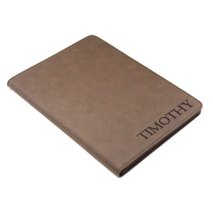May include: A rectangular brown portfolio case with a smooth, matte finish and stitched edges. The name TIMOTHY is engraved in a dark, serif font on the lower right corner of the front cover. The case is closed and features a zipper closure.
