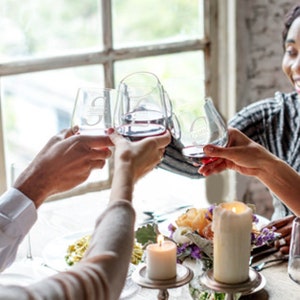 May include: Three clear stemless wine glasses filled with red wine, each featuring a unique etched monogram letter and a name. The glasses are held up in a toast, showcasing the personalized engraving on the glass surface against a bright window.