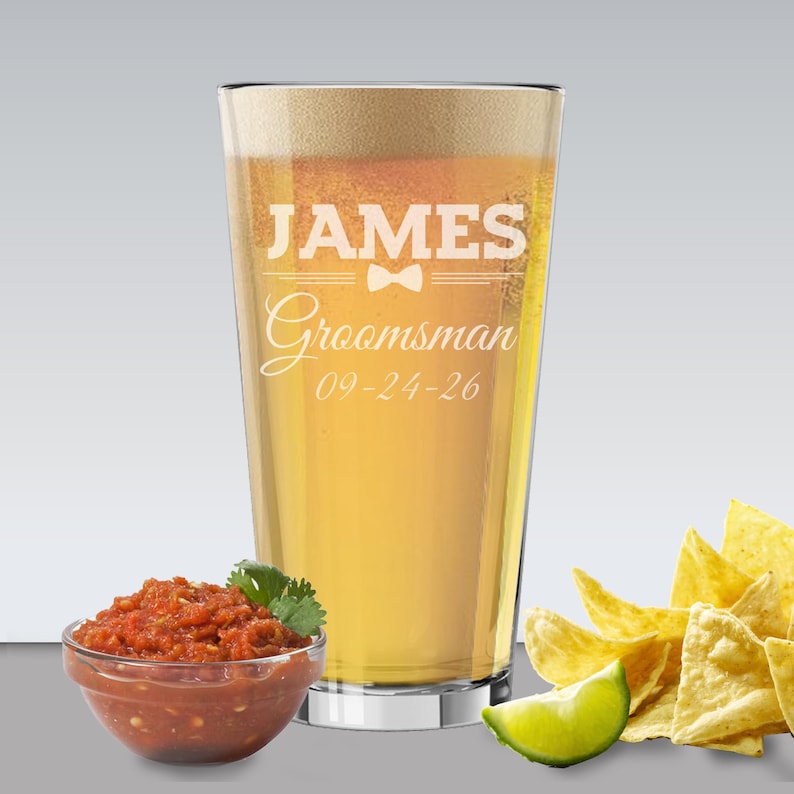 May include: A pint glass filled with beer, featuring the text "JAMES Groomsman 09-24-26" in white. A small bowl of salsa, tortilla chips, and a lime wedge are in the foreground. The glass has a silver base.