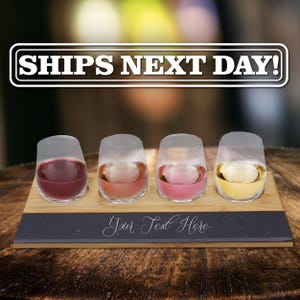 May include: A rectangular bamboo serving board with four circular cutouts holding stemless glass tumblers filled with different colored liquids. A dark gray slate inlay at the front features the white cursive text Your Text Here.