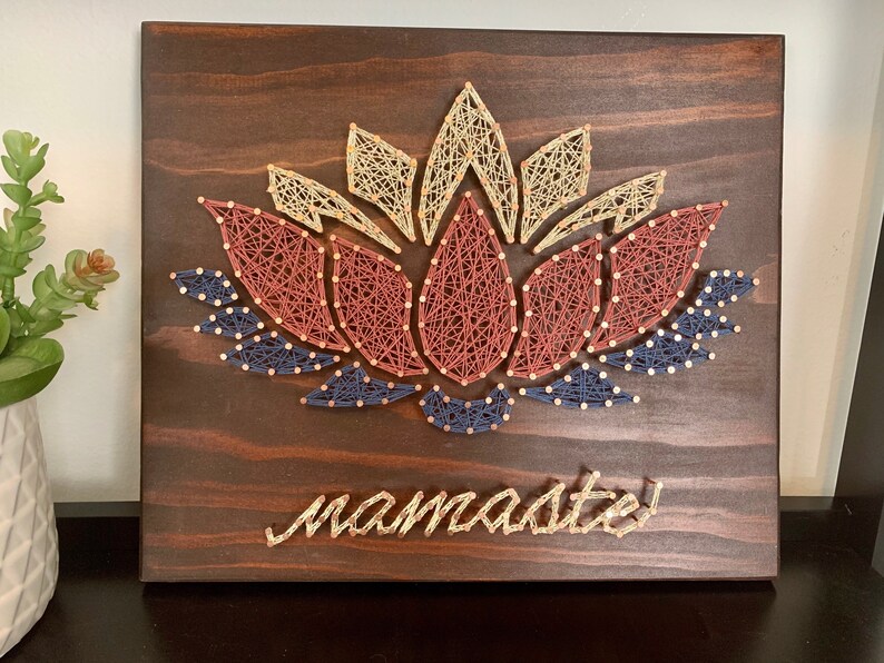 May include: A wooden wall hanging with a string art design of a lotus flower in shades of red, blue, and beige. The word "namaste" is written in gold string art below the flower.