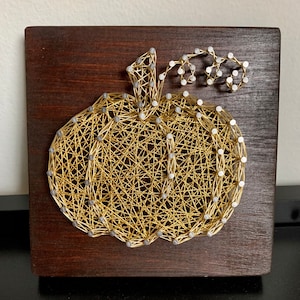 May include: A gold string art pumpkin with a silver stem and vine on a dark brown wooden background.