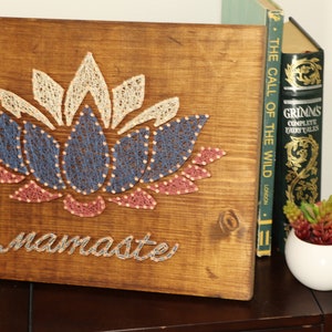 May include: A wooden wall hanging with a string art design of a lotus flower in shades of pink, blue, and white. The word "namaste" is written below the flower.