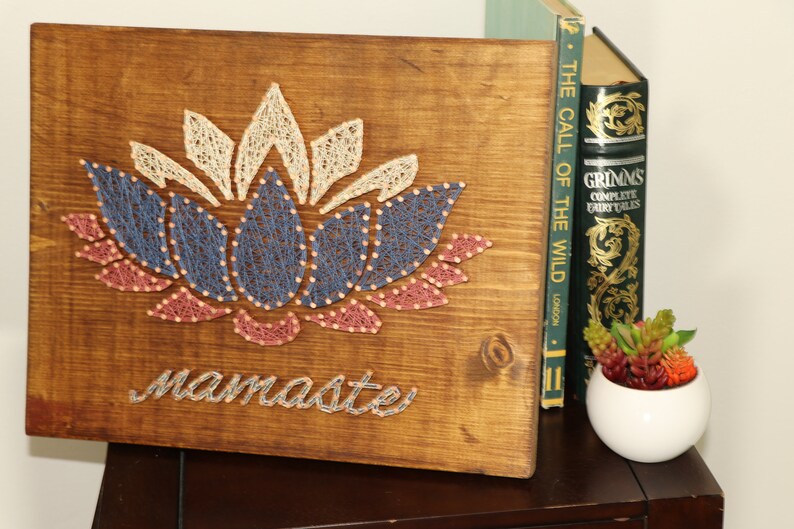 May include: A wooden wall hanging with a string art design of a lotus flower in shades of pink, blue, and white. The word "namaste" is written below the flower.