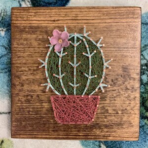 Op de afbeelding: Een string art cactus met een roze bloem erop, in een roze pot, op een houten achtergrond.