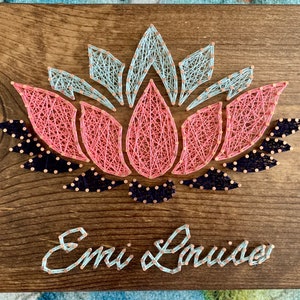 May include: A wooden sign with a pink, blue, and black string art lotus flower design. The name "Emi & Louise" is written below the flower in blue string.