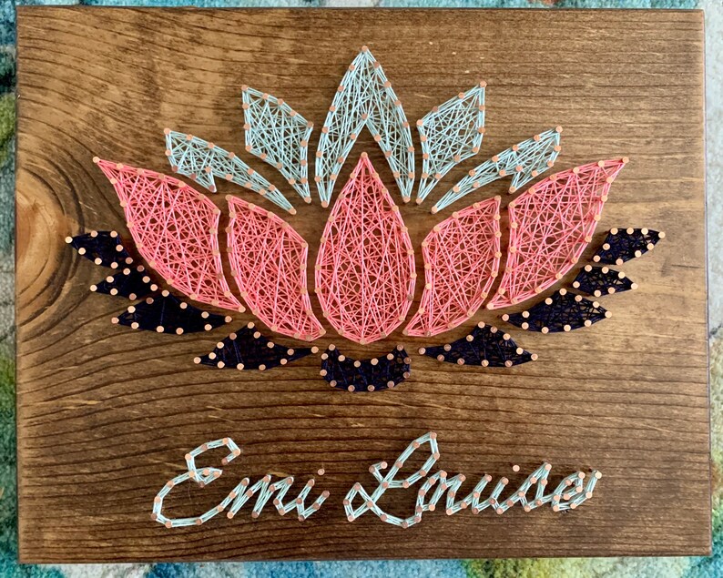 May include: A wooden sign with a pink, blue, and black string art lotus flower design. The name "Emi & Louise" is written below the flower in blue string.