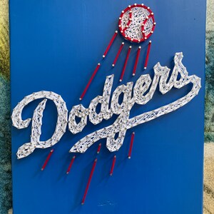 May include: A blue wooden sign featuring the Los Angeles Dodgers logo, crafted with white string and red accents. The baseball and the word "Dodgers" are meticulously designed with string art, creating a unique and eye-catching piece of wall decor.