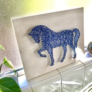 May include: Blue string art of a horse on a white wooden background. The horse is outlined with nails and strung with blue thread.