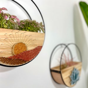 Op de afbeelding: Twee houten wandplanters met zwarte metalen hoepels. De planter aan de linkerkant heeft een string art zonsondergangontwerp met roze en groene planten. De planter aan de rechterkant heeft een blauw en groen string art ontwerp met een roze plant.