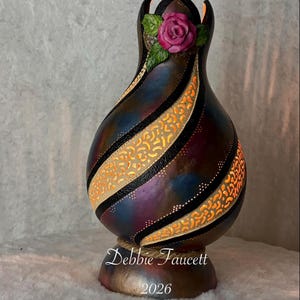 May include: A decorative gourd lamp with a rose accent. The lamp features a swirling design with black stripes and cut-out patterns that emit a warm glow. The base is a mix of brown and gold, with the text "Debbie Faucett 2026".