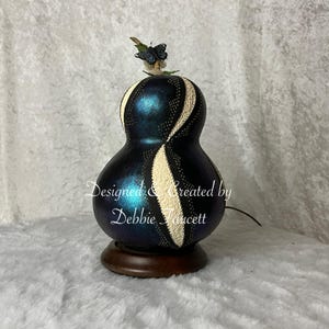 May include: A decorative gourd lamp with a dark blue iridescent finish and cream-colored, patterned accents. A small butterfly adorns the top. The lamp sits on a round, wooden base. Text on the gourd reads "Designed & Created by Debbie Faucett."