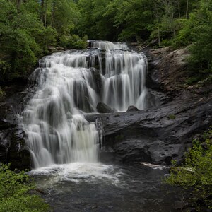 May include: A scenic photograph of a cascading waterfall. The water flows over dark, layered rocks, creating a contrast with the white, flowing water. Lush green trees and foliage surround the waterfall, adding depth and a natural feel to the landscape.