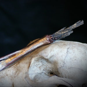 May include: A long, thin, copper-coloured wand with a black, frayed tip. The wand is resting on a white, textured surface.
