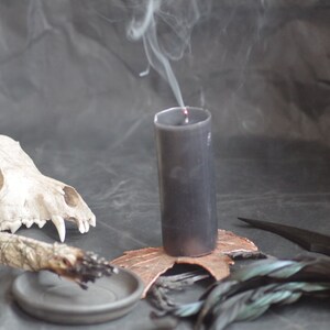 May include: A black candle with smoke rising, next to a dog skull, a bundle of sage, a small dish, a copper leaf, and a feather. The items are arranged on a dark surface, creating a mystical or spiritual theme.