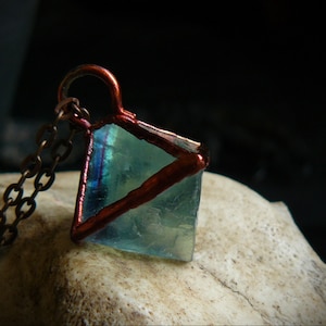 Puede incluir: Un colgante envuelto en alambre de cobre con un cristal de fluorita azul claro. El colgante está unido a una cadena de bronce.