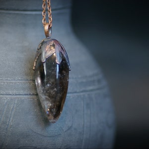 Puede incluir: Un colgante de cristal marrón oscuro con un engaste y una cadena de cobre. El cristal tiene forma de lágrima y tiene un ligero brillo.