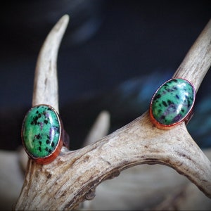 May include: Two oval-shaped rings with green and black gemstone settings. The rings are made of copper and are set on a light brown antler.