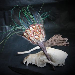 Puede incluir: Un abanico decorativo de plumas con plumas marrones, beige y verde azulado, un mango marrón oscuro y largos acentos verdes. También hay un cráneo de animal blanco, un manojo de hilo marrón y un cuchillo negro.